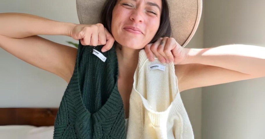 woman wearing a tan hat with her eyes closed and smiling holding up a dark green women's sweater and a beige women's sweater