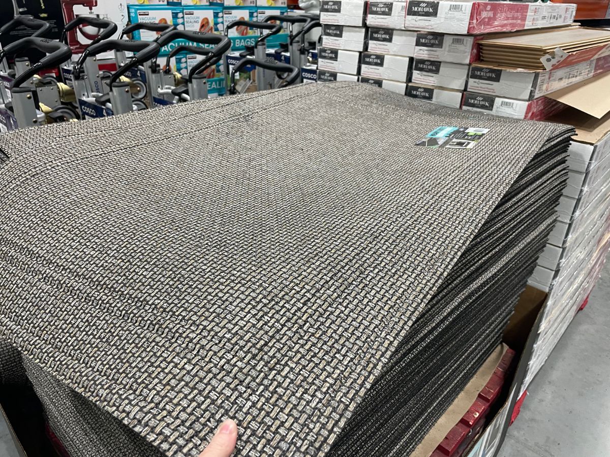 a womans hand displaying the texture of a heavy duty door mat stacked on a pallet in a costco club