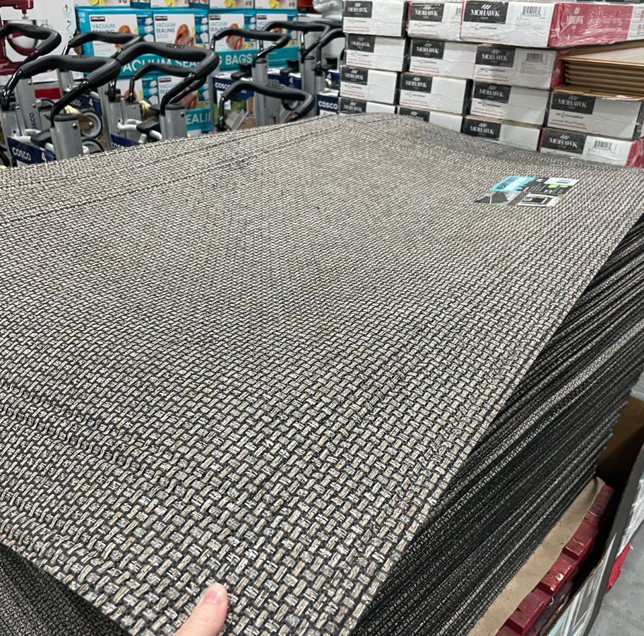 a womans hand displaying the texture of a heavy duty door mat stacked on a pallet in a costco club