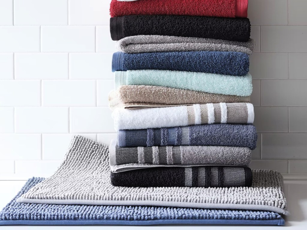 Large stack of JCPenney, bath towels, on top of bathmats in front of white tile wall
