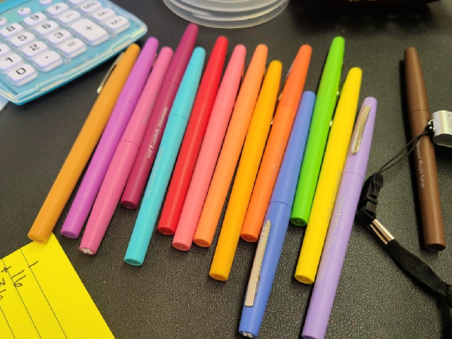 Different colored Paper Mate flair pens on desktop next to keyboard and notebook