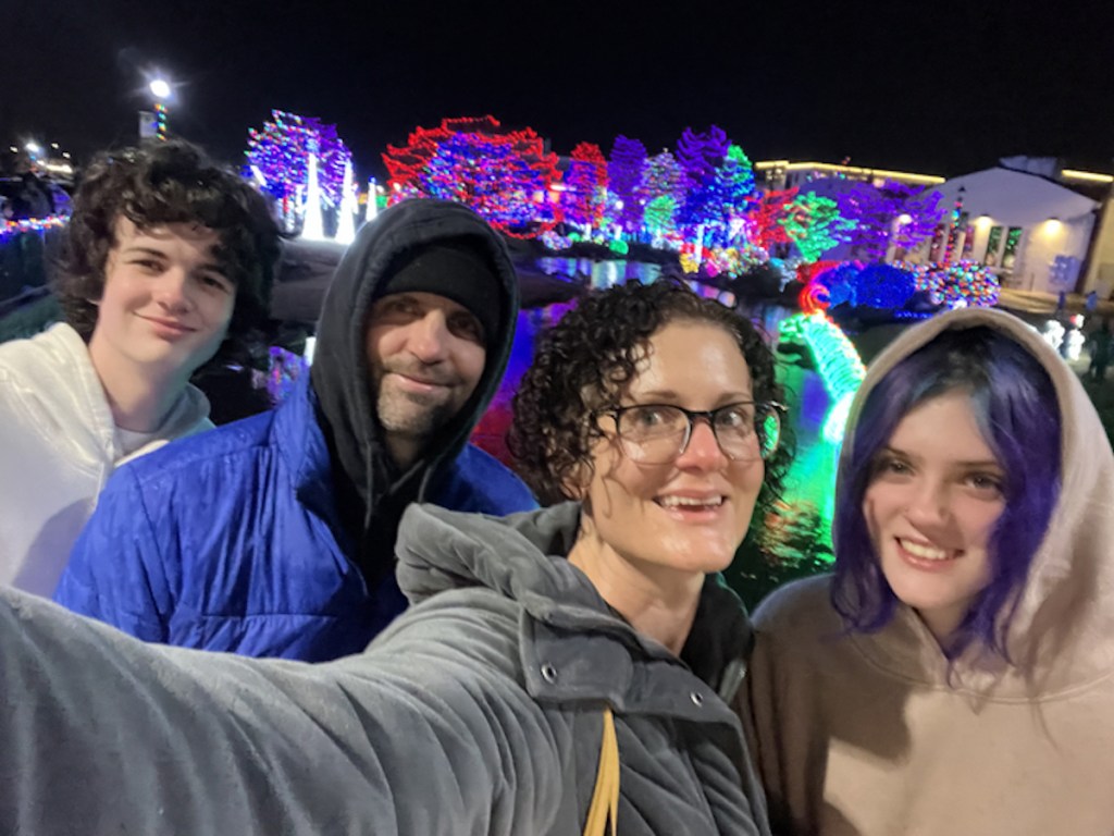 family taking selfie outside on rainy night in front of house covered in colorful christmas lights