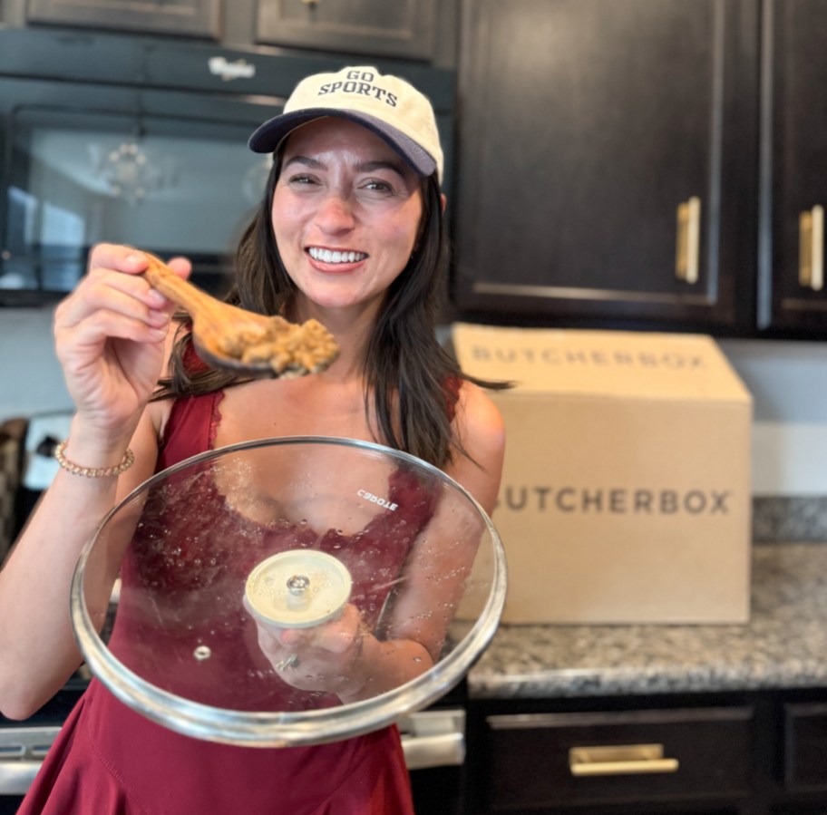 smiling woman holding a cooking spoonful of meat towards the camera, above a pan lid