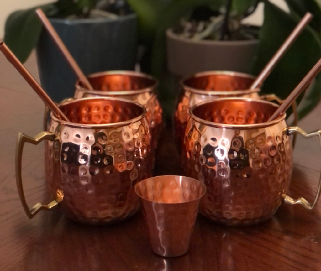 Set of gold copper mugs with matching straws sitting on wood table in front of plant
