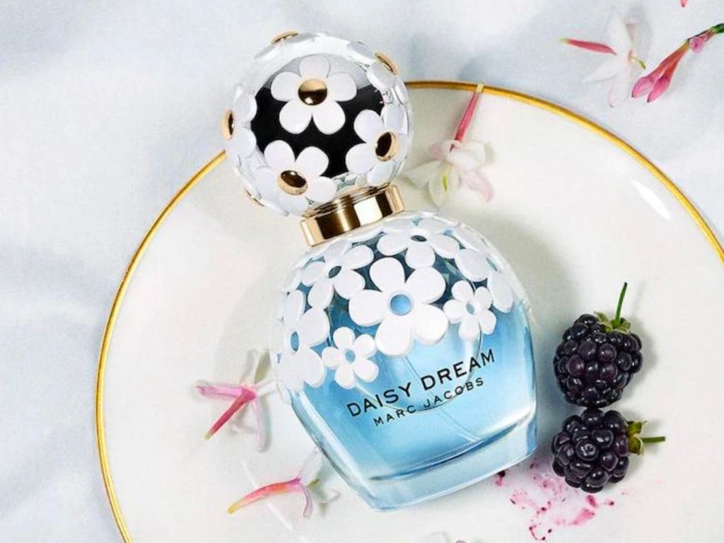 a 1 ounce bottle of daisy dream by marc jacobs on a gold rimmed china saucer next to 2 black berries