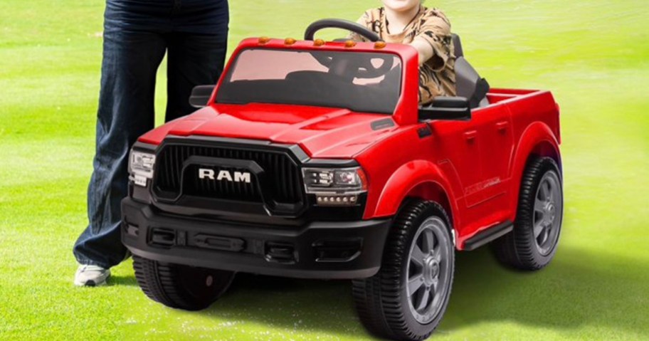 child riding in red dodge ram ride on car in grass with parent next to it 