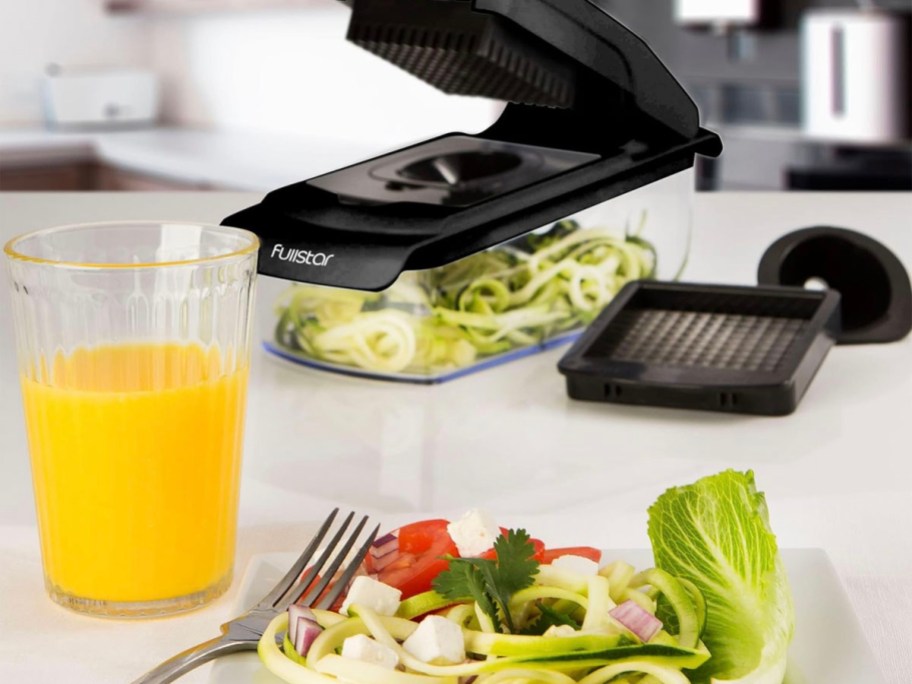 black and clear vegetable chopper on countertop with cup of orange juice and plate of veggies