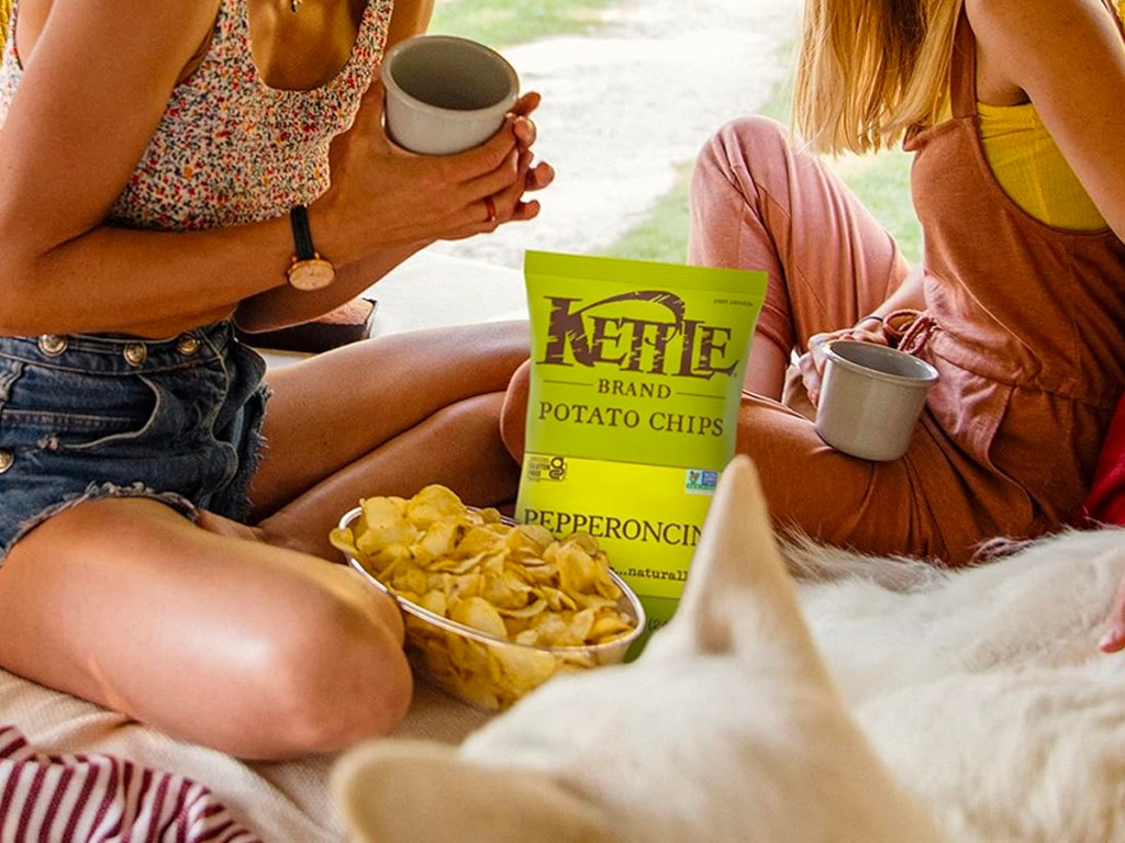 two women sitting with a bag of kettle peppercini chips in a bowl next to bag