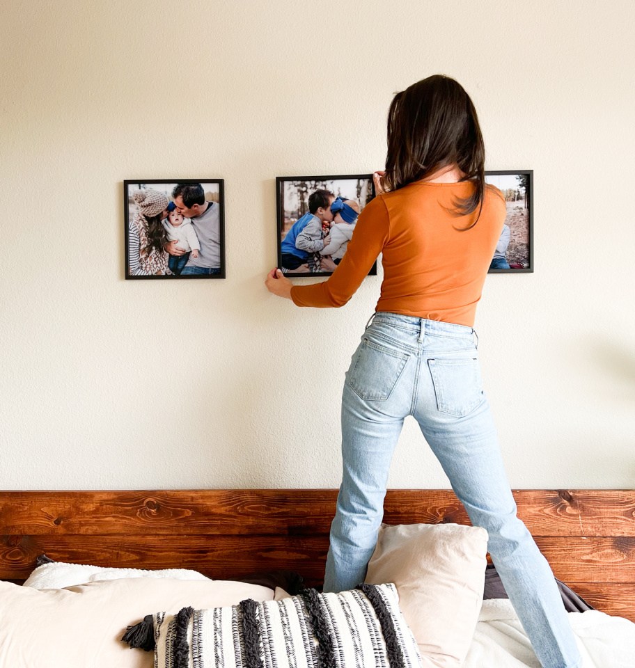 woman standing on bed hanging black tilepix frames on walls