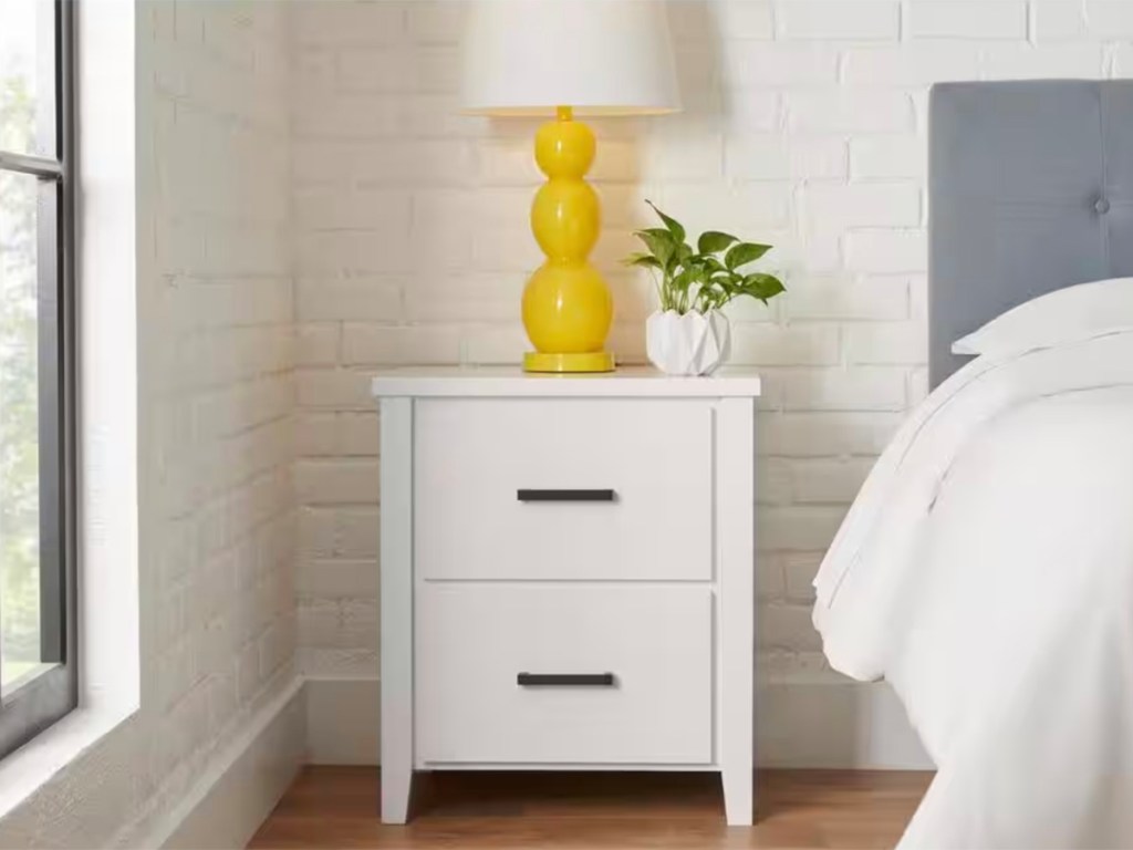 white 2 drawer nitstand next to bed with yellow lamp on top