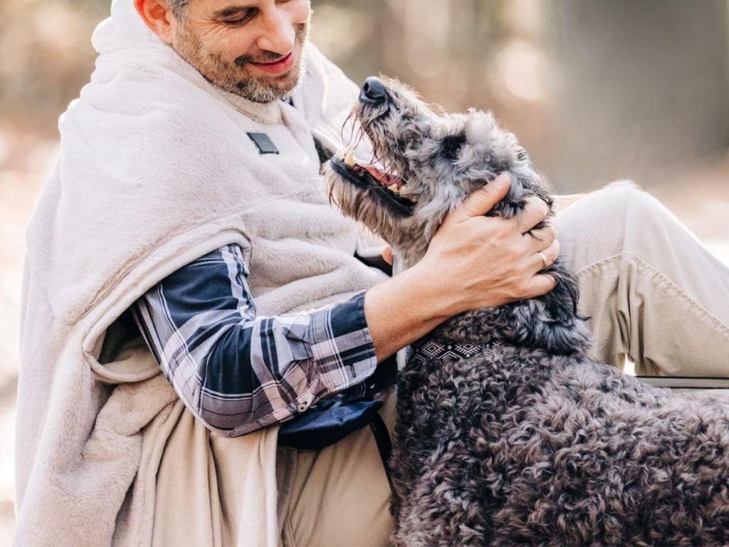 man wearing a heated blanket petting a dog
