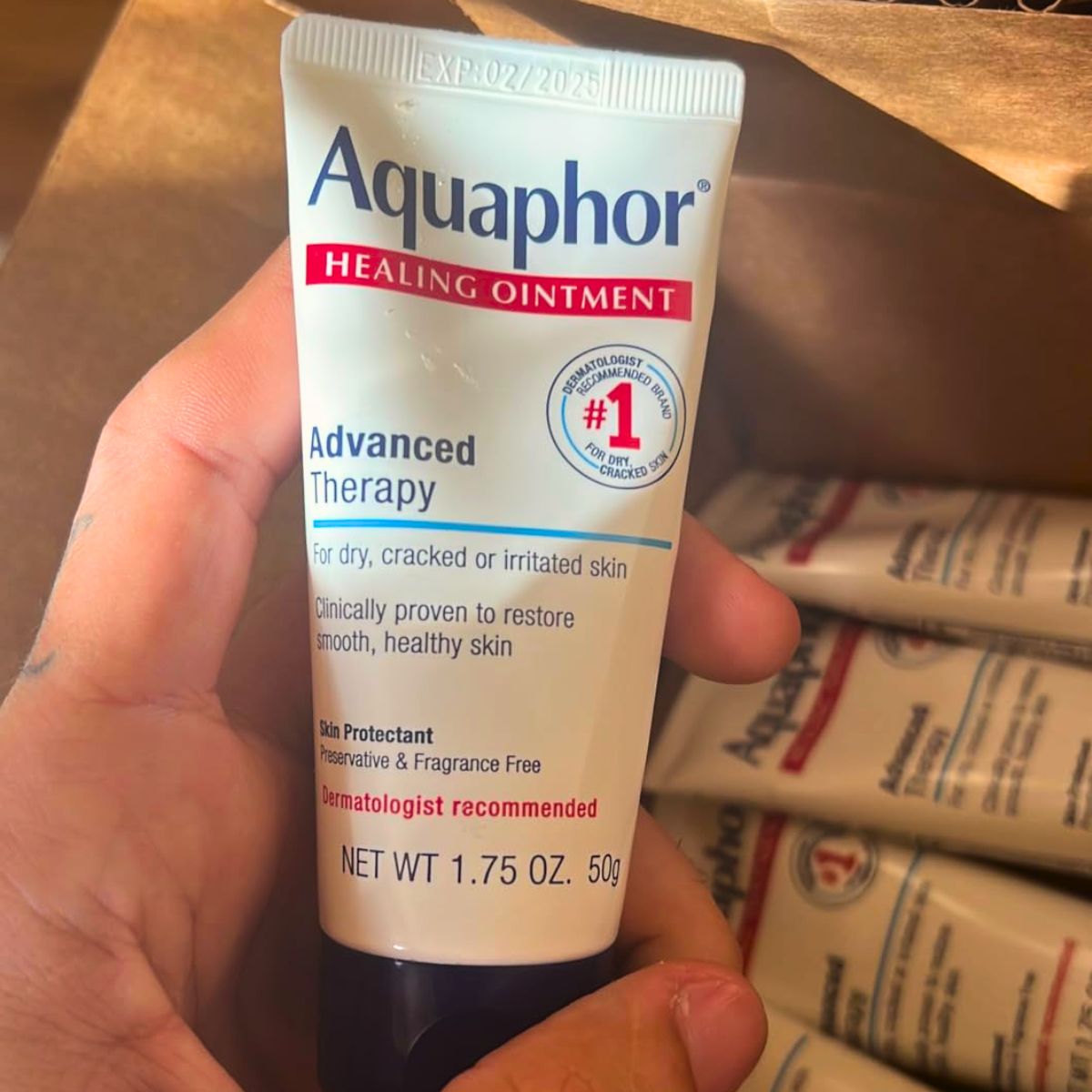 a hand holding a tube of Aquaphor Advanced Therapy Healing Ointment with the rest of 6 pack in background