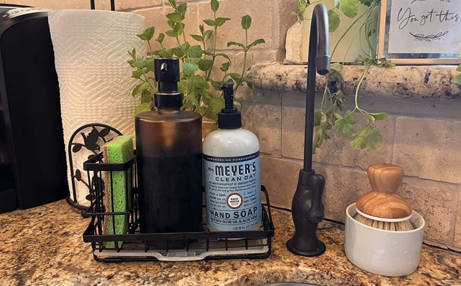 a black steel sink caddy with sponge, hand soap and dish soap on a granite counter by a sink