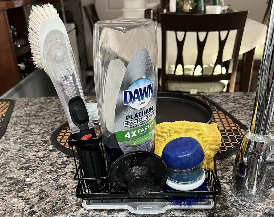 a black steel sink caddy with scrub brush, drain stoppers, dish liquid and scrub daddy on a granite counter by a sink