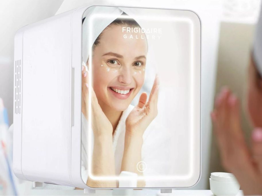 A woman looking into a small refrigerator with a mirror on it 