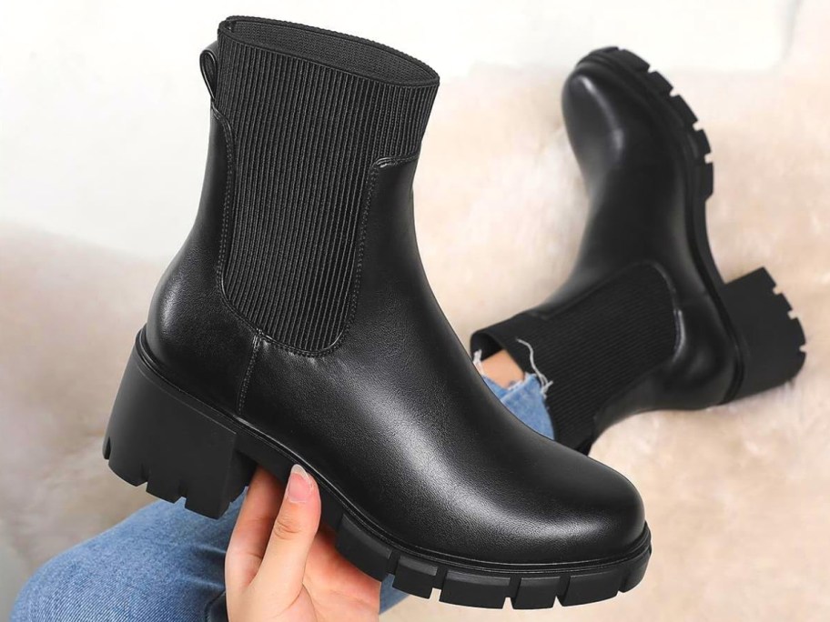 hand holding a black lug sole women's Chelsea boot, wearing the other on her foot in the background