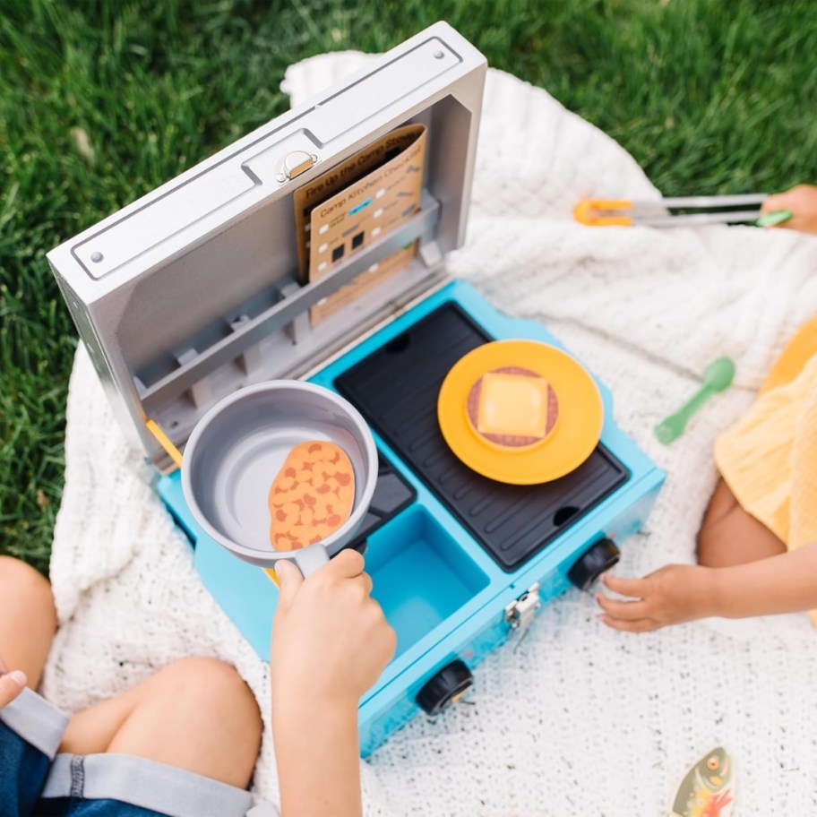 kids sitting on a blanket on the grass playing with a toy camp stove and food set