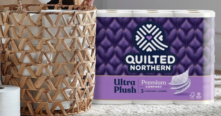 pack of 12 Quilted Northern Ultra Plush Premium Comfort Toilet Paper Mega rolls sitting beside a tan basket with more rolls and a roll on the floor