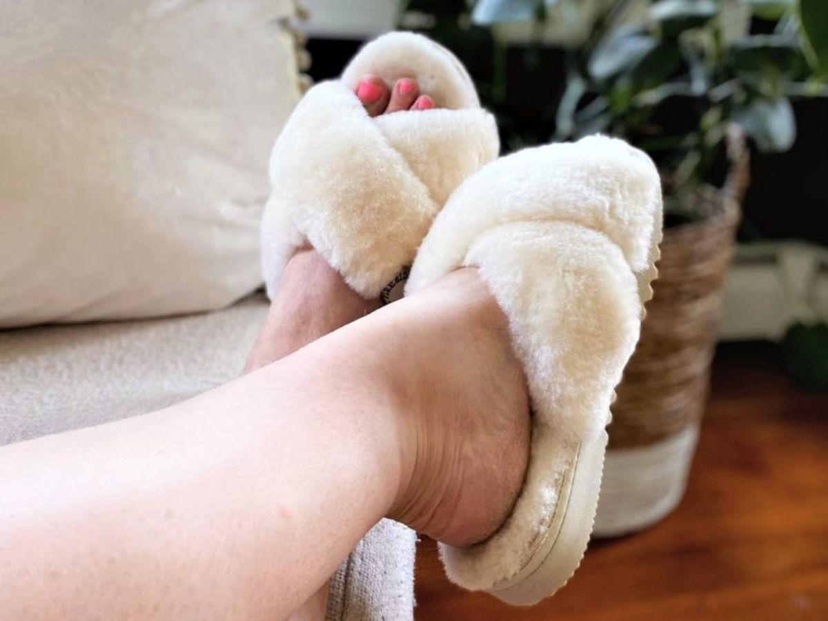 woman's legs and feet sitting on a sofa wearing a cream color pair of furry Dearfoams slippers