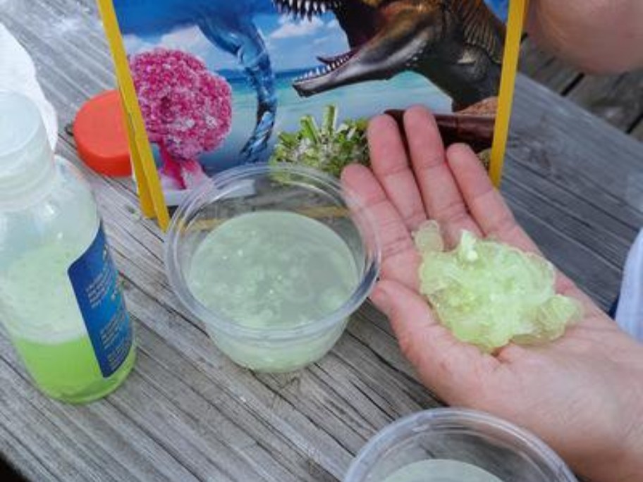 kids hand holding a yellow crystal grown from a science experiment, with containers of liquid and other items, and the box the scient experiments came in