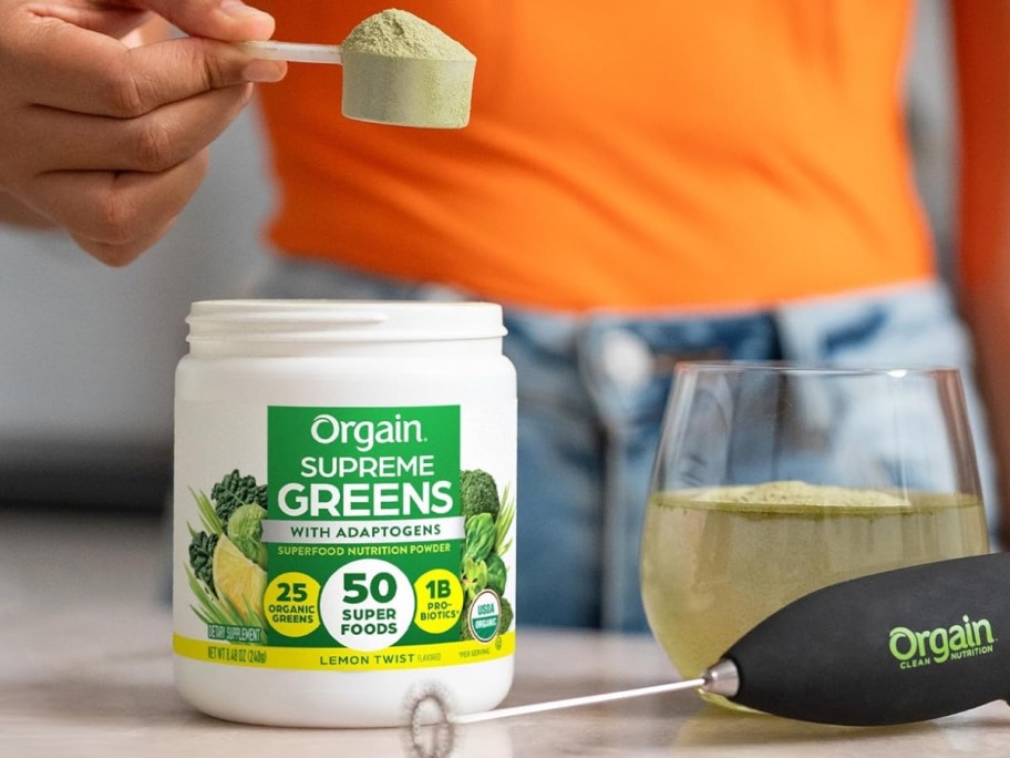 container of Orgain Supreme Greens Powder open with person getting a scoop of it to add to a glass of water