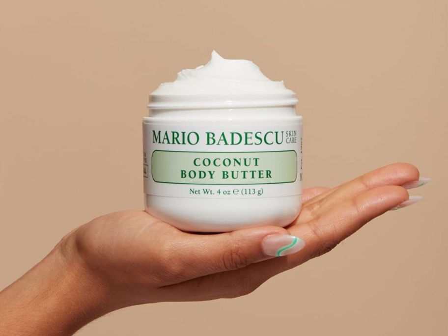 A hand holding an open jar of Mario Badescu Coconut Body Butter