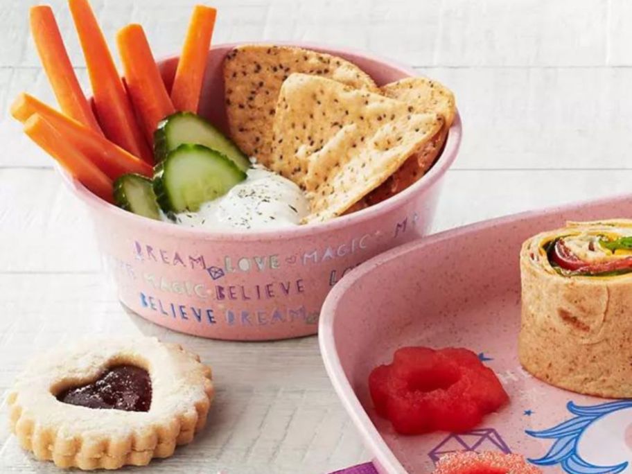 Close up view of Member's Mark kids dinnerware bowl filled with veggies, dip, and tortilla chips and a plate with watermelon cutouts and a roll-up sandwich on it