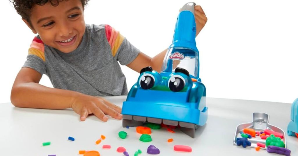 a little boy at a table pushing a small blue plastic vacuum to pick up pieces of play-doh