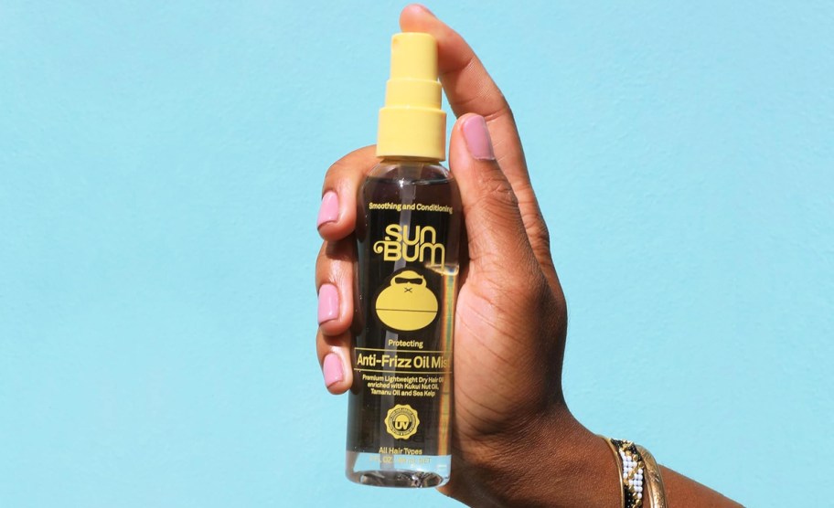 hand holding a bottle of Sun Bum Anti-Frizz Oil Mist with blue background