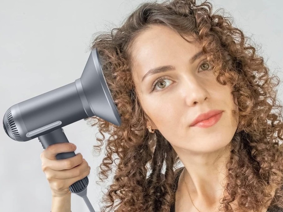 woman holding up grey hair dryer with diffuser attachment to her curly hair