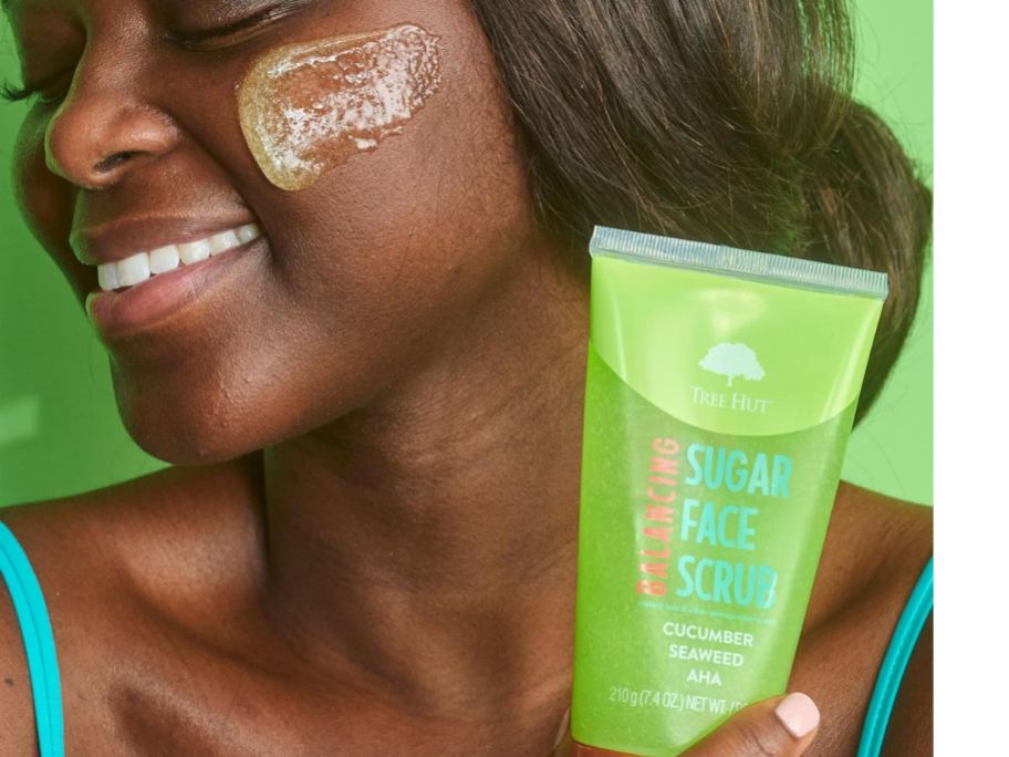 woman holding a tube of Tree Hut Balancing Sugar Face Scrub with some of the scrub on her cheek