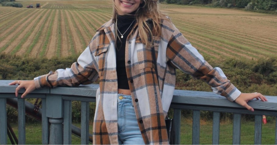 woman wearing a brown flannel shacket with a black crop top and jeans with her arms stretched on a back deck, field behind her