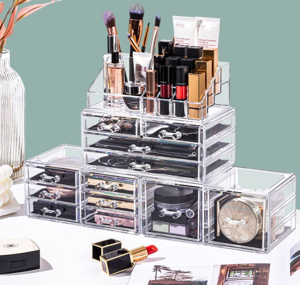 a four piece acrylic jewelry organizer stacked in 3 tiers and filled with a range of cosmetics, from lipstick tubes to eyeshadow palettes next to a vase of flowers and sitting on a white counter top