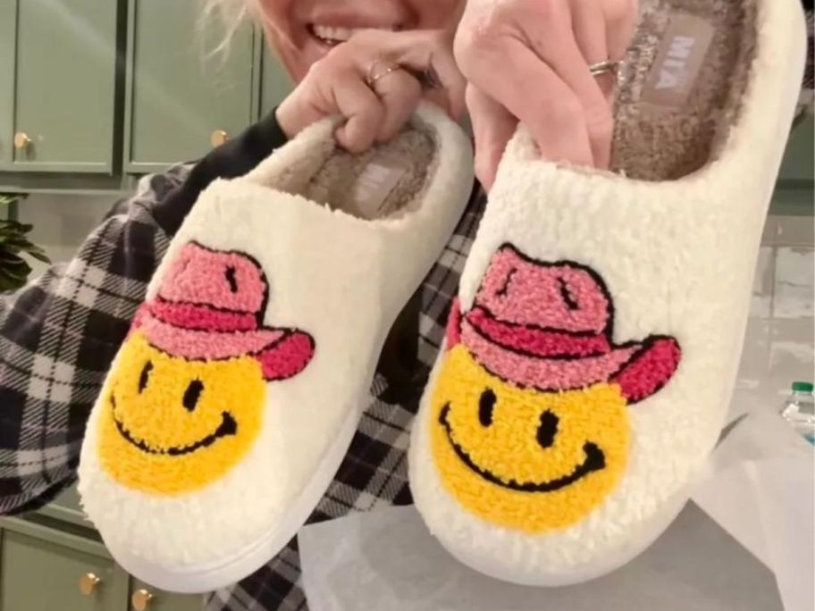 woman holding up a cute pair of Mia slippers with smiley faces wearing pink cowboy hats on them