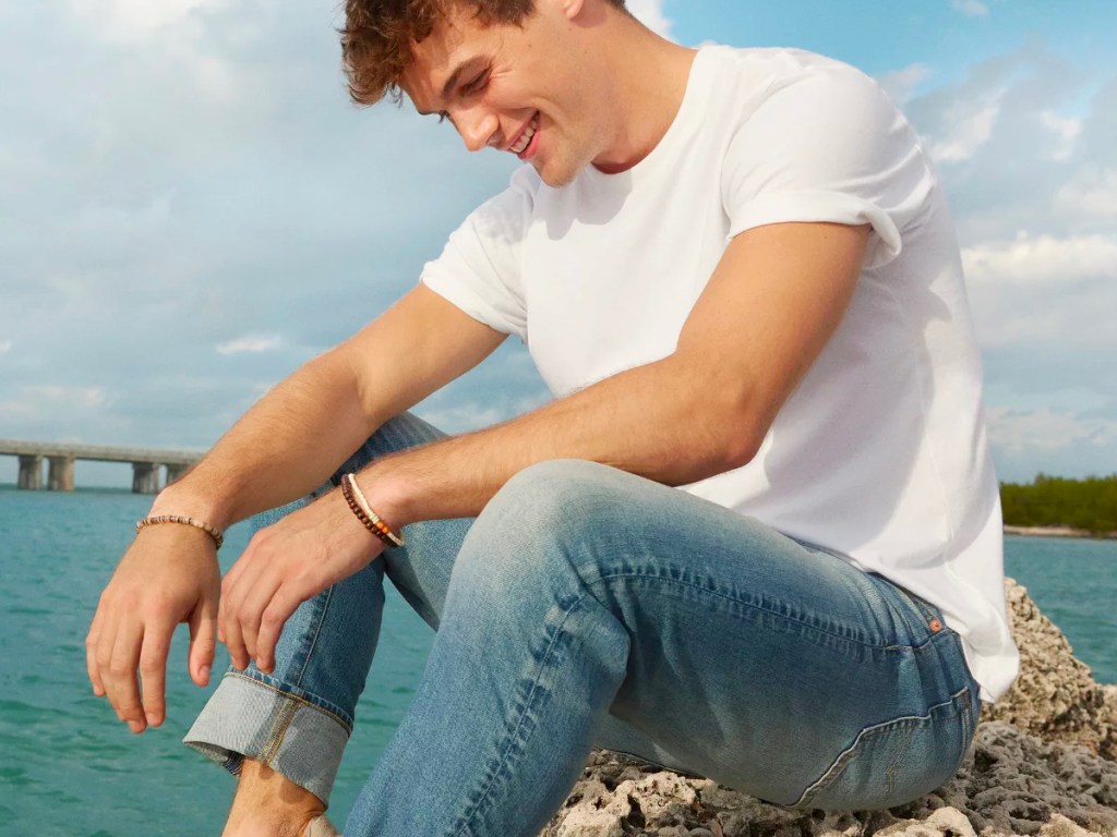 man wearing white tee and jeans on rocks next to water