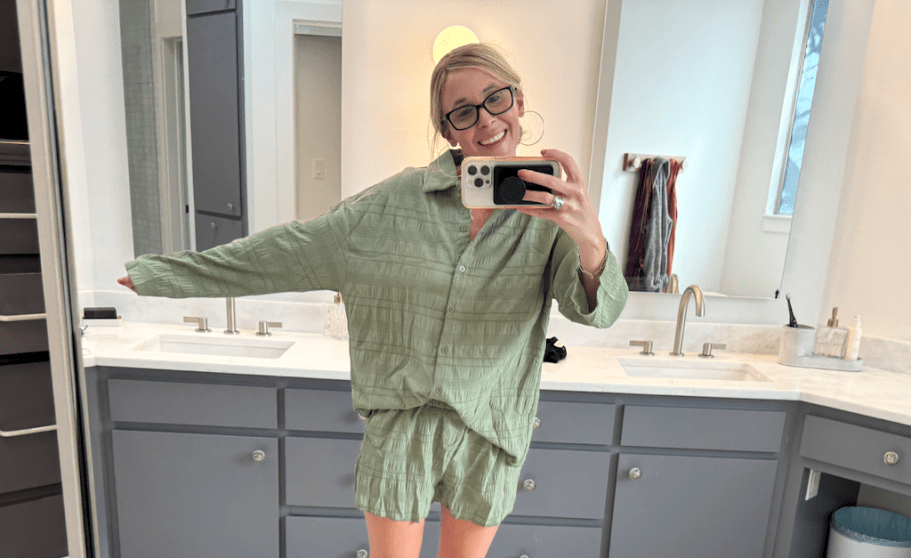 woman taking photo of herself wearing long sleeve green shirt and shorts in bathroom
