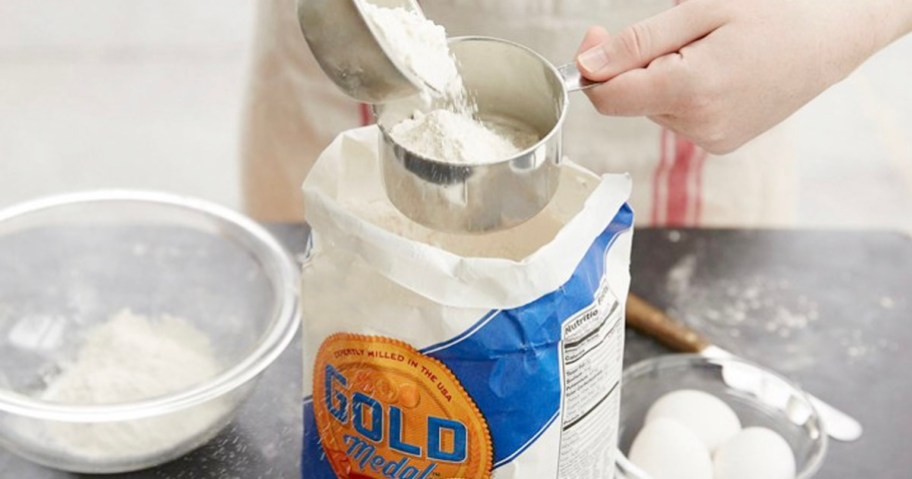 person taking flour out of bag with measuring cup sitting next to bowl of flour