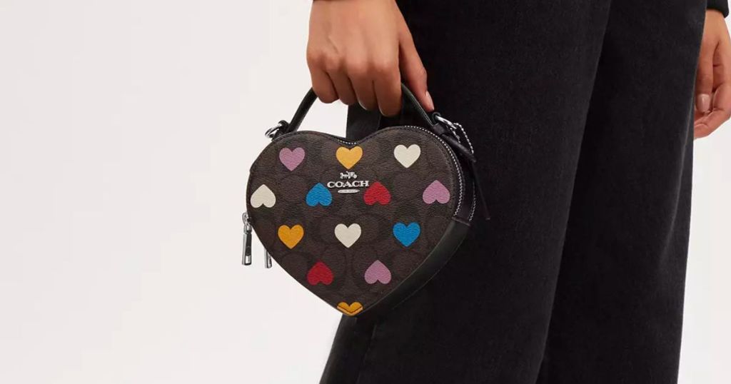 a womans hand holding a small heart shaped coach bag
