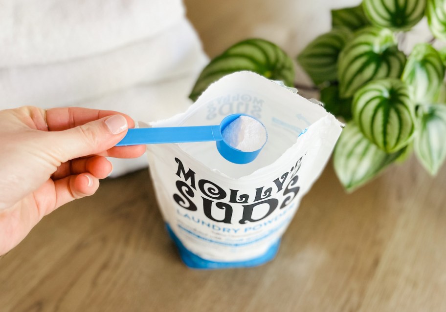 hand scooping powder laundry detergent from bag on wood table with towels and plant