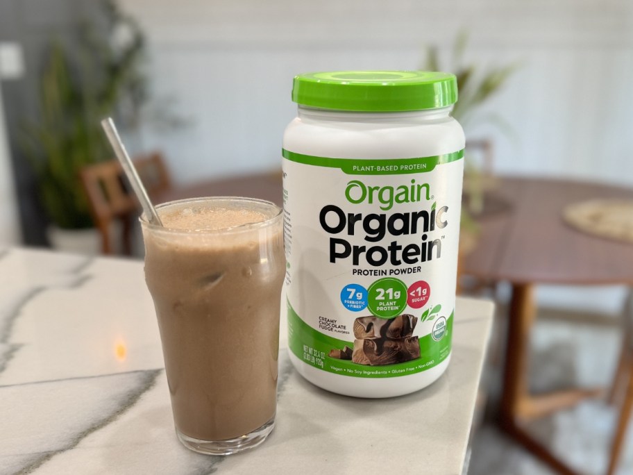 large container of orgain protein powder on kitchen counter next to glass of it mixed up