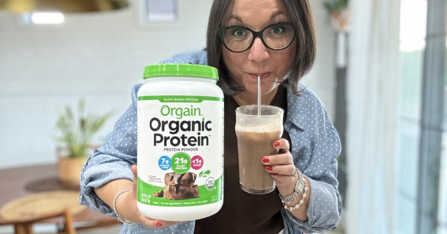 Lina (woman) holding a large canister of Orgain Protein Powder while sipping on a glass of the chocolate protein shake
