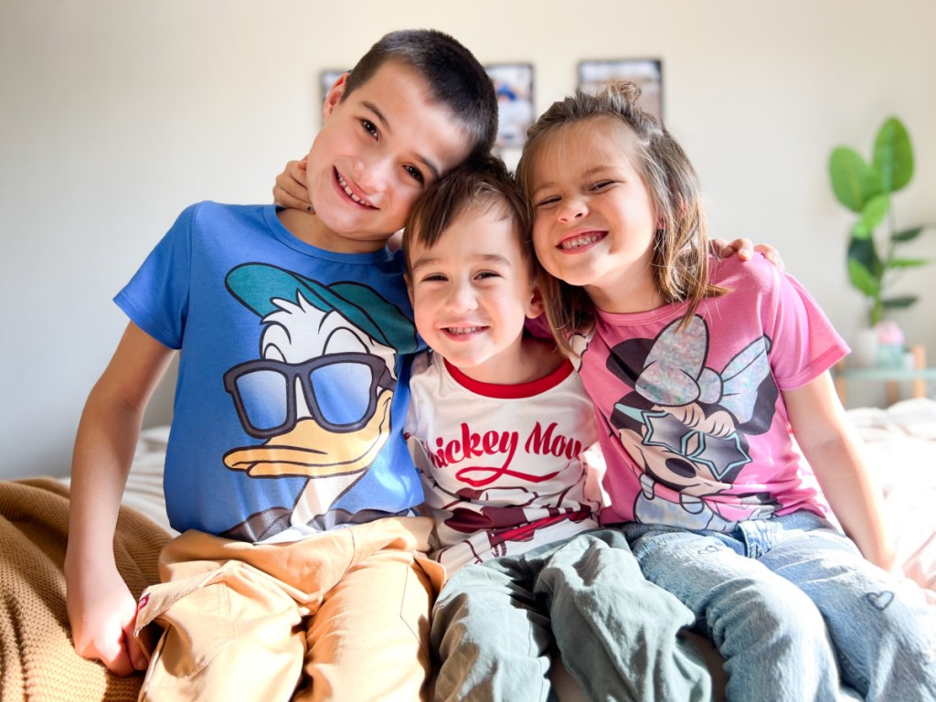 three smiling kids sitting on the end of a bed, wearing patpat disney shirts