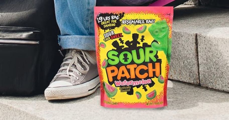 a family size bag of watermelon sour patch kids candy sitting on step next to a girl's foot