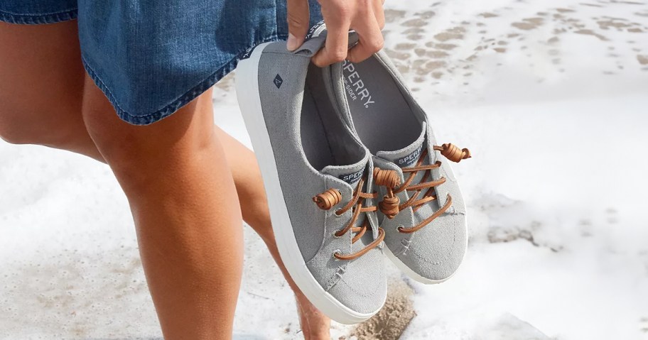 woman holding gray sperry shoes while walking in beach water