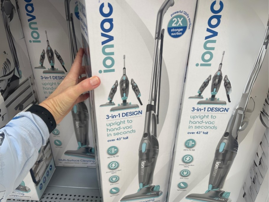 hand raching for a box with a IonVac 3-in-1 Corded Stick Vacuum on a shelf with more of the vacuums