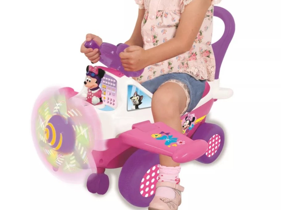 little girl riding a pink, white, and purple Minnie Mouse airplane ride on toy