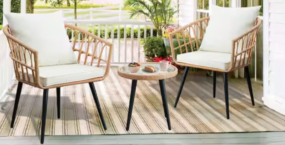 Cozy porch scene with two wicker chairs with white cushions and a small round table. On the table are a coffee cup and pastries. Lush greenery in the background.