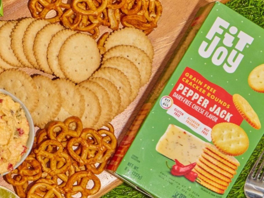 A charcutterie board with crackers and pretzels on it next to a box of Fit Joy Pepperjack Crackers