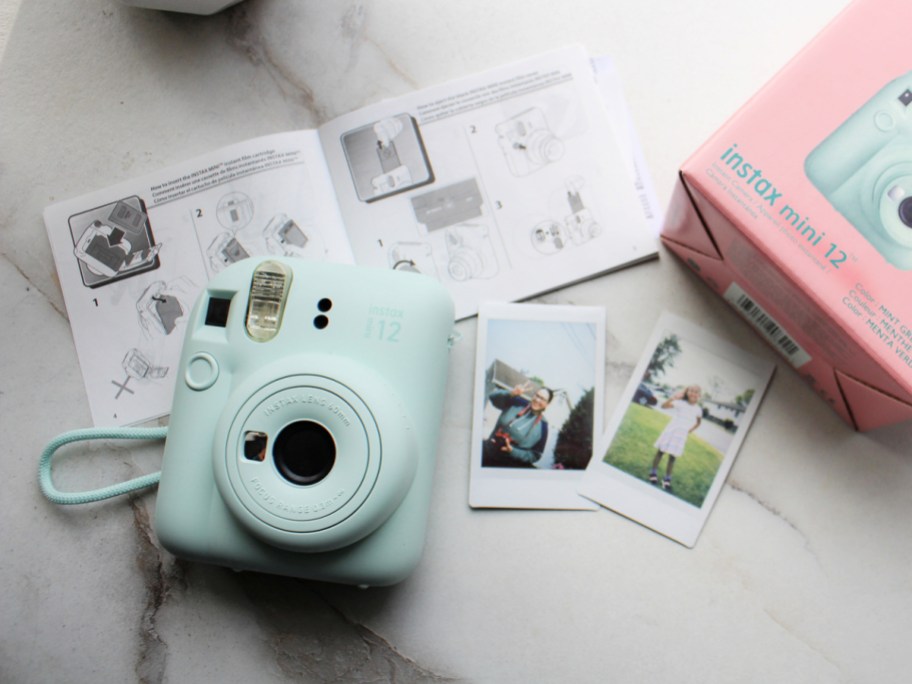Fujifilm Instax Mini 12 camera next to an instruction guide, photos and packaging