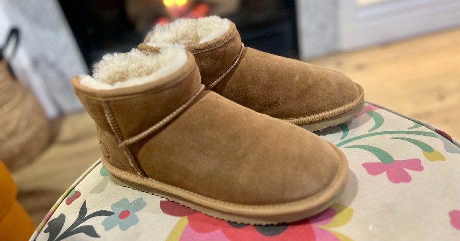 a pair of women's tan booties with shearling lining that look like UGGS sitting on an ottoman in a living room, a fireplace is in the background